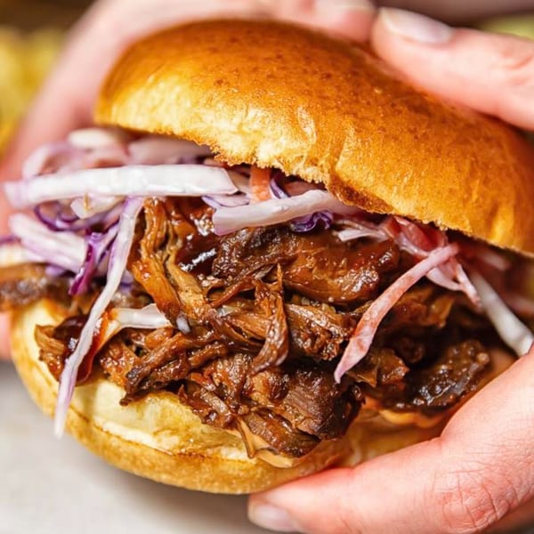Hands holding a pulled pork sandwich with coleslaw on a brioche bun at Riley's Place.