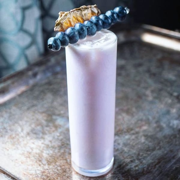 Tall glass of light purple drink garnished with blueberries on a skewer and dried pineapple.