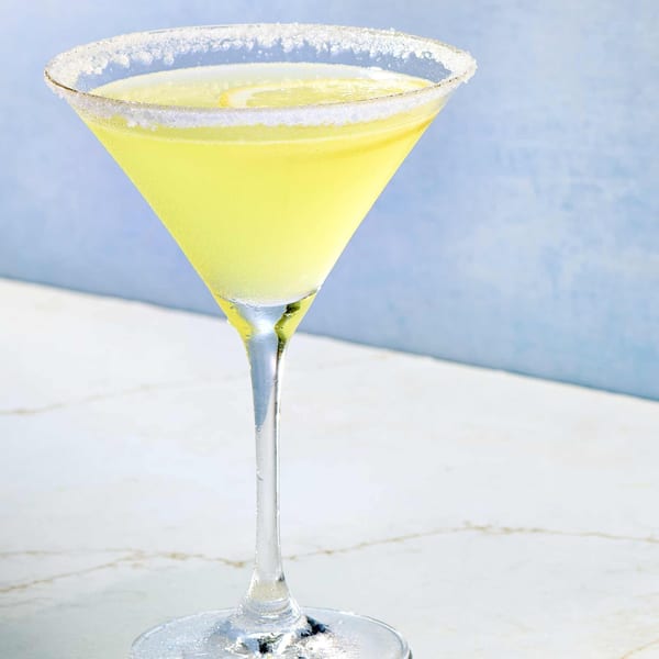 A vibrant yellow martini in a sugar-rimmed glass, with a lemon slice, on a marble surface.