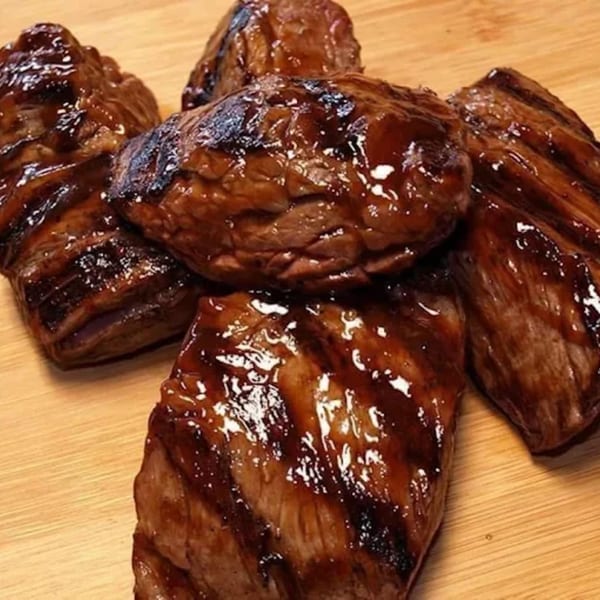 Five pieces of glistening, dark brown grilled meat, possibly steak or ribs, on a light wooden surface.