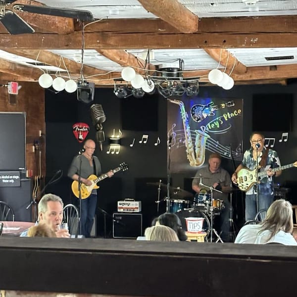 A band performs live on stage at Riley's Place, with musicians playing guitar and drums.