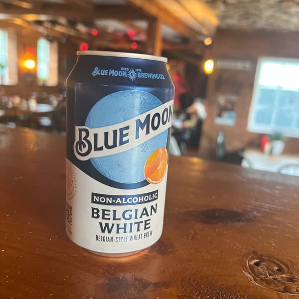 A Blue Moon non-alcoholic Belgian White beer can sits on a wooden bar at Riley's Place.