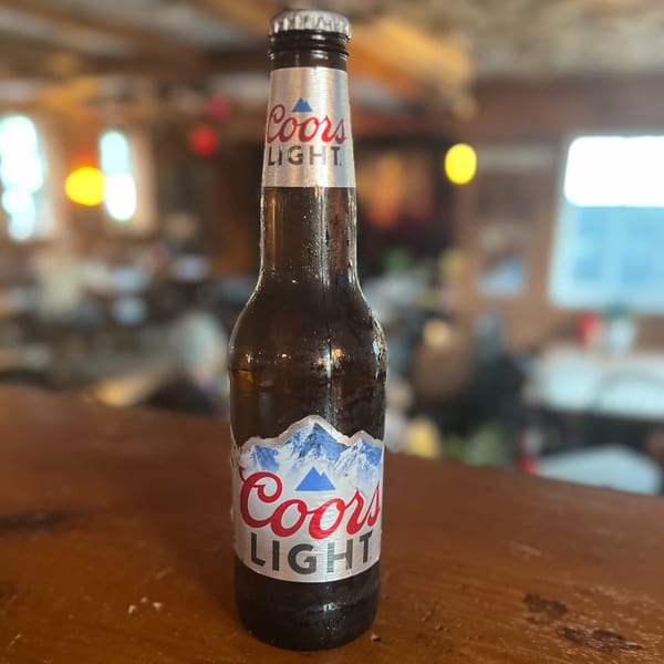 Sweaty Coors Light bottle on a wooden bar top with a blurred restaurant interior in the background.