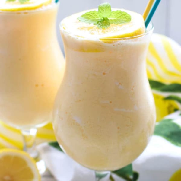 Two glasses of creamy yellow lemon smoothies, garnished with lemon slices and mint, with straws.