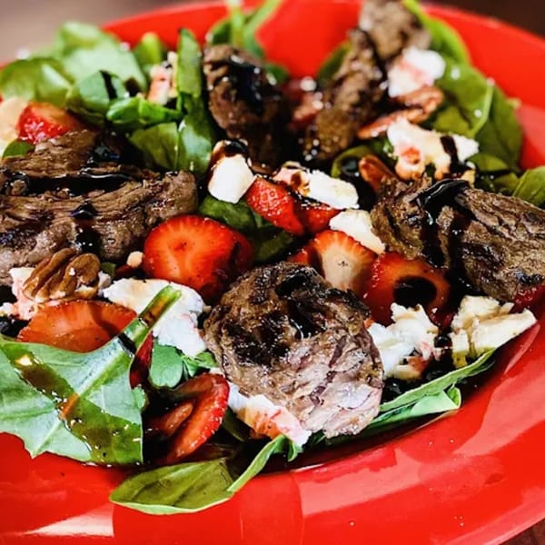 A vibrant salad with steak, strawberries, pecans, and feta cheese, drizzled with balsamic glaze.