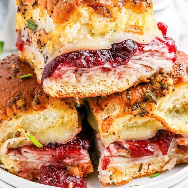 Stacked turkey and cranberry sliders with melted cheese and poppy seeds on a white plate.