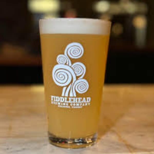 A glass of Fiddlehead Brewing Company beer with a foamy head sits on a wooden surface.