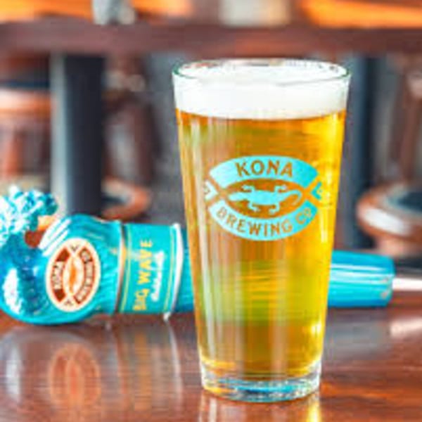 A pint glass of Kona Brewing Co. beer stands on a wooden table next to a blue bottle.