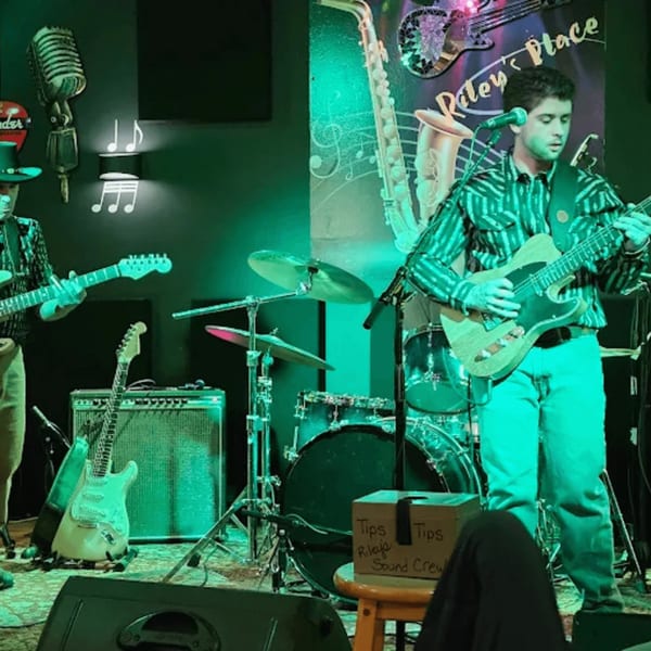 Two musicians playing electric guitars on a stage with drums and a "Riley's Place" sign.