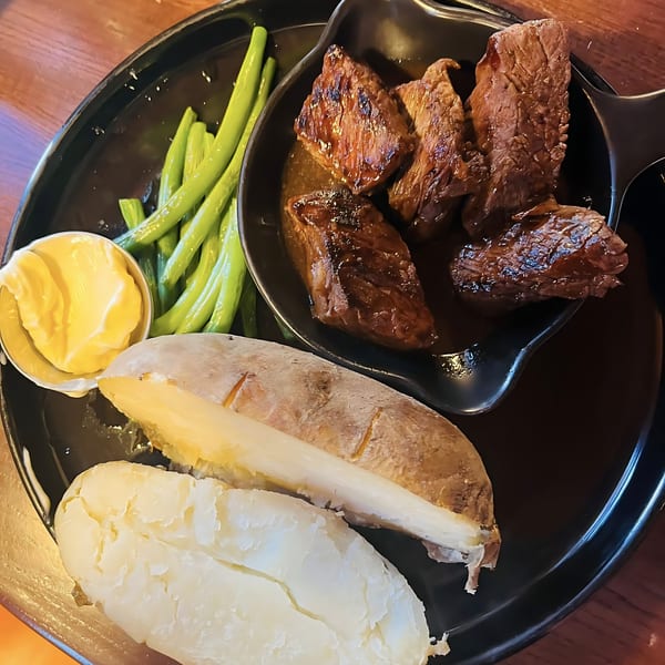 Grilled steak bites in sauce, green beans, and a baked potato with butter on a black plate.