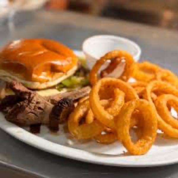 A roast beef sandwich with curly fries and a side of sauce on a white plate.