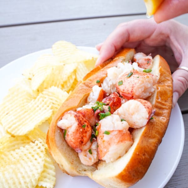 A hand squeezes lemon over a lobster roll with waffle-cut chips on a white plate.