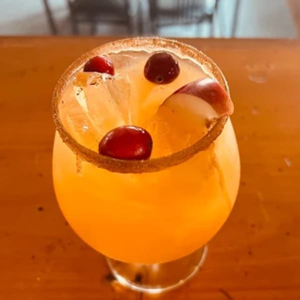 A vibrant orange cocktail with cranberries and apple slices, rimmed with cinnamon sugar.