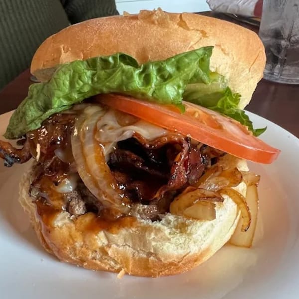 A close-up of a burger from Riley's Place, topped with caramelized onions, bacon, cheese, lettuce, and tomato.