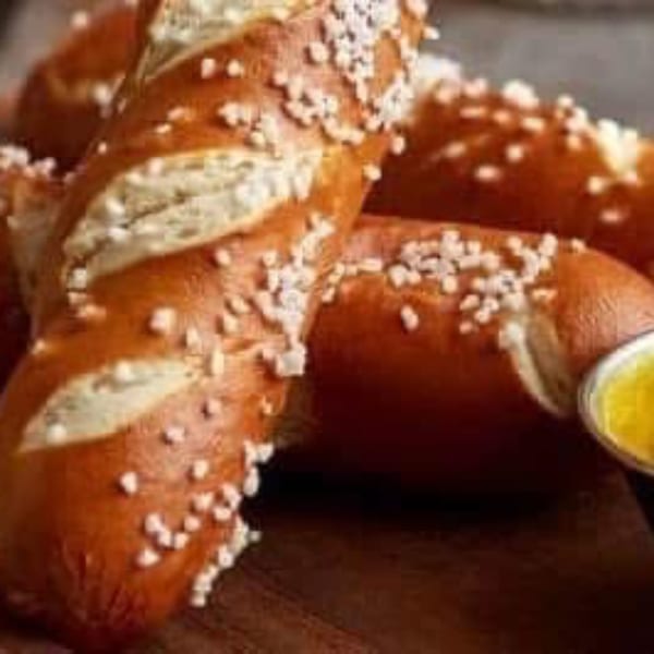 Close-up of golden-brown pretzel sticks, generously sprinkled with coarse salt, next to a small cup of melted butter.