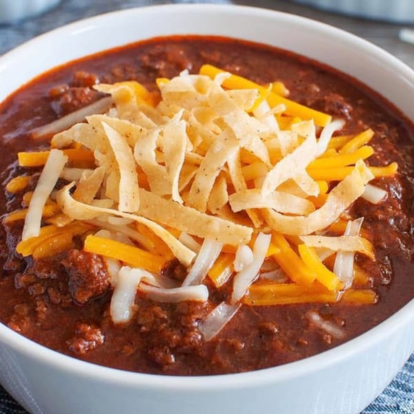 A white bowl of Riley's Place chili topped with shredded cheddar, white cheese, and tortilla strips.