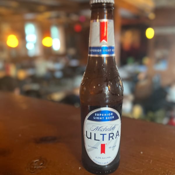 A Michelob Ultra beer bottle, condensation on its surface, sits on a wooden bar at Riley's Place.
