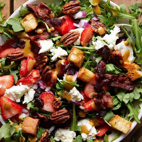 A vibrant salad with arugula, strawberries, pecans, bacon, feta, croutons, and dressing.