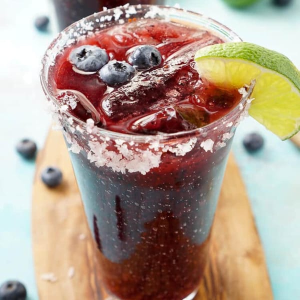 A blueberry margarita with a salted rim and lime wedge, on a wooden board.