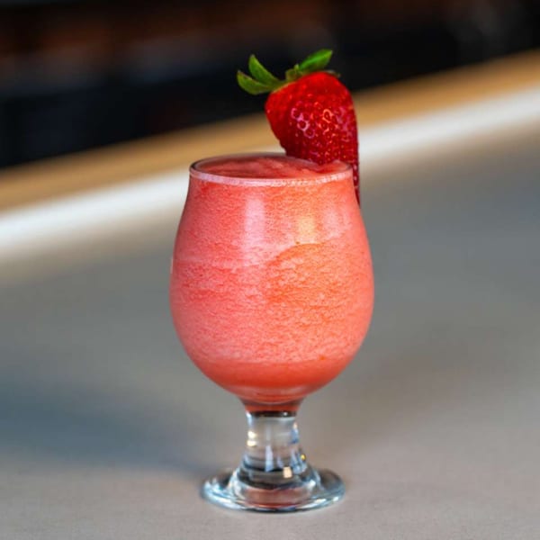 A vibrant red frozen drink, garnished with a fresh strawberry, sits on a light-colored bar.